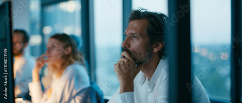 Focused man in a modern office setting, deep in thought while working on a computer with colleagues in the background, evening light creating a contemplative atmosphere.