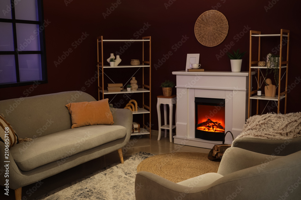 Obraz premium Cozy living room interior with a lit electric fireplace, comfortable sofa, and decorative shelves at night