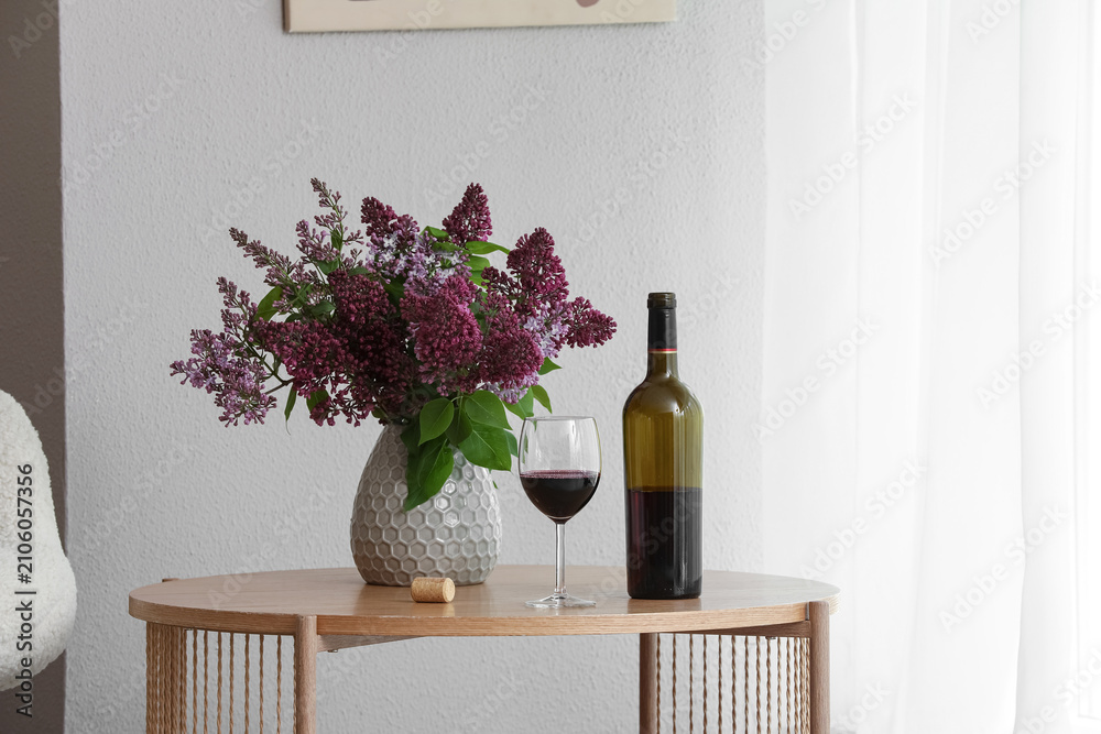 Obraz premium Red wine bottle and glass with a vase of purple lilac flowers on a wooden table indoors, Set indoors with a white wall and a sheer white curtain in the background, illuminated by natural light