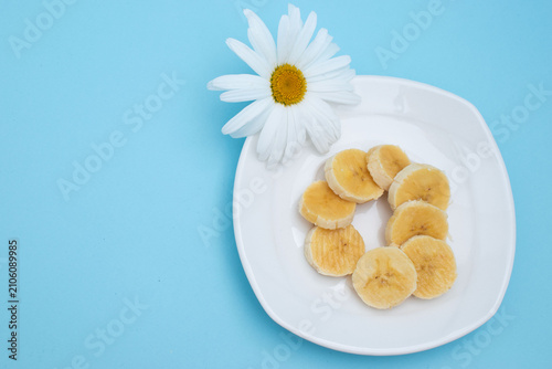 Banana slices on white plate with daisy flower on blue background