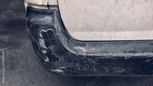 Close up of a heavily damaged black plastic car bumper with severely cracked, peeling, and flaking paint, showing deep scratches and weathered texture on a white vehicle.