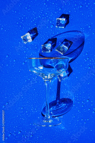 Empty cocktail glass with ice cubes on a wet, vibrant blue summer background.