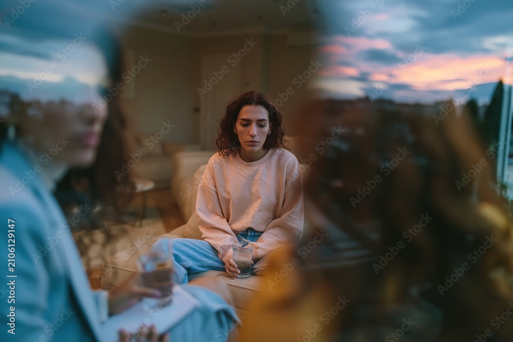 Obraz premium A sad young woman holding a glass of water during therapy, reflecting on her mental health in a soft pink and blue room for psychotherapy and wellbeing
