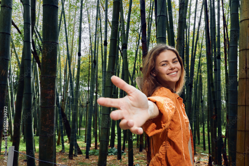 Fototapeta premium Woman in bamboo forest reaches hand toward camera, smiling in orange jacket. Portrait of young woman connecting with nature, walkway among tall green bamboo stalks.