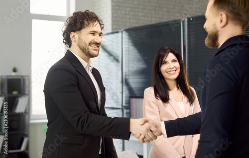 Businessmen handshake at meeting in office. A smiling colleague observes as partners confirm agreement and show strong teamwork. Clear message of trust, partnership, and business success.