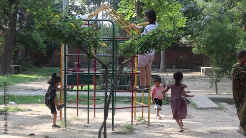 A grandmother carries a little child while walking through a village playground, as several children play together on outdoor equipment nearby.