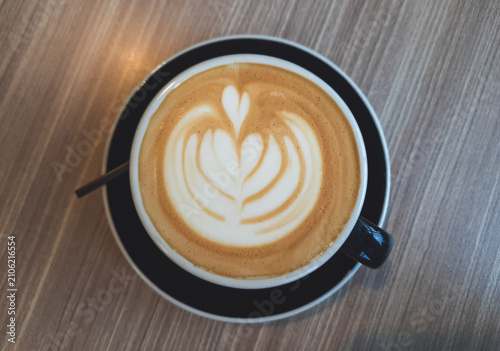 Warm latte art blooms in creamy coffee. Black cup rests on wooden table. Soft light highlights delicate foam design. Spoon lies beside cup, ready for use. Cozy, quiet moment captured for comfort