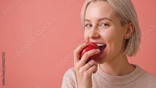 A young, blonde woman in a knit sweater smiles while taking a bite of a red apple against a pink background.