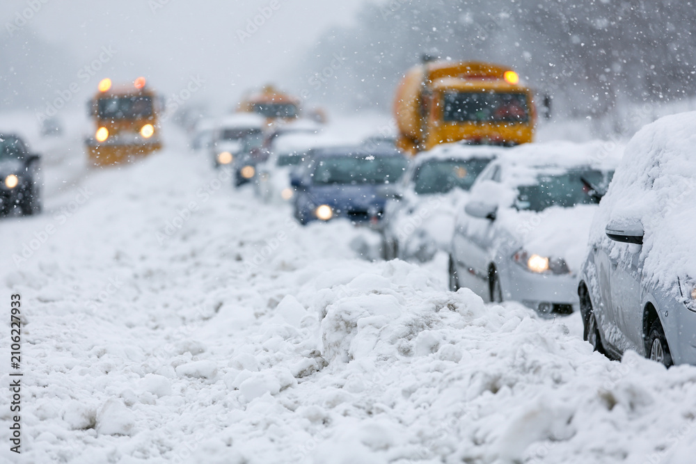 Obraz premium Snow blankets roads, turning streets into white, icy paths. Vehicles halt, lights dimmed as snowflakes drift downward. Yellow trucks wait patiently amid the quiet chaos