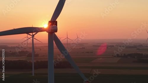 A beautiful view of a wind farm during golden hour, with the sun shining in a bright orange sky and multiple wind turbines silhouetted against the field.
