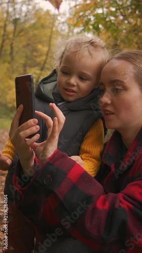 Mother and toddler sharing a warm autumn moment outdoors while looking at a smartphone together in a colorful forest filled with golden seasonal foliage