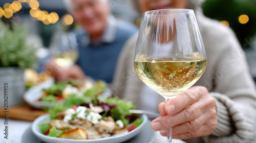 Closeup of elderly woman holding wine glass over plated salad during alfresco lunch for senior dining program, healthful meal and community socializing