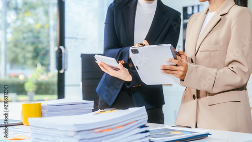 Two business women standing in an office reviewing reports with a tablet and phone beside a stack of papers on a desk