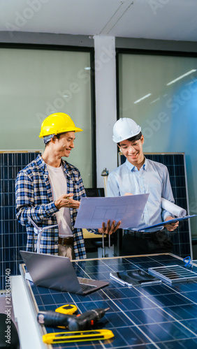 Two engineers wearing hard hats discuss solar panel plans in an office while reviewing documents and a laptop