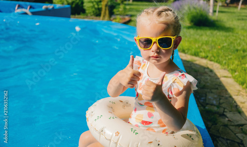 Cute girl in sunglasses with swim ring showing thumbs up by swimming pool.