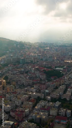 Vertical video. Alanya, Turkey. Aerial drone flight over dense residential rooftops toward cloud-covered Taurus Mountains, with sun rays breaking through above a sandy Mediterranean beach. Aerial Vie
