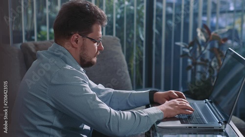 Man working on laptop at home sitting near window typing on keyboard and completing online tasks of remote work in calm home environment