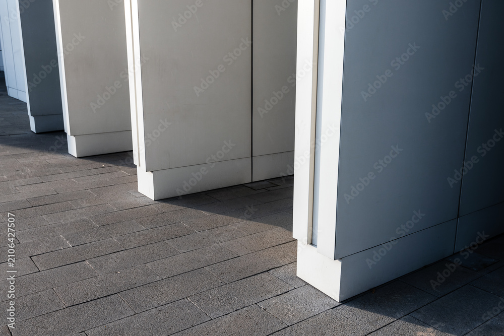 Obraz premium Minimalist modern building exterior with a row of white architectural columns casting long geometric shadows across textured urban pavement, evoking contemporary design and abstraction