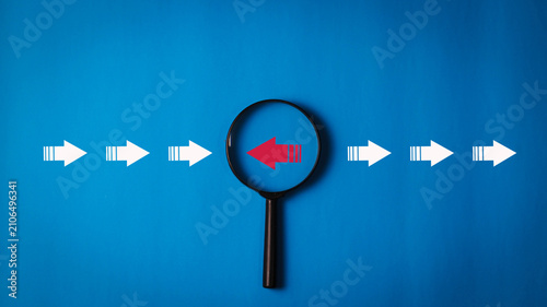 Fotografía Magnifying glass highlighting a red arrow pointing opposite to white arrows on a blue background, symbolizing change, disruption, independent thinking, leadership, business, and decision making