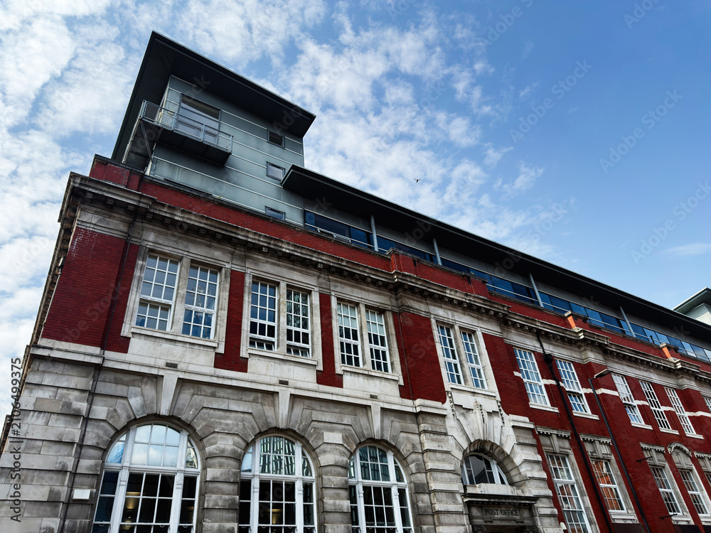 Fototapeta premium Historic red brick post office building with modern rooftop extension in Manchester, UK, urban architecture and ornate stone facade under a blue cloudy sky