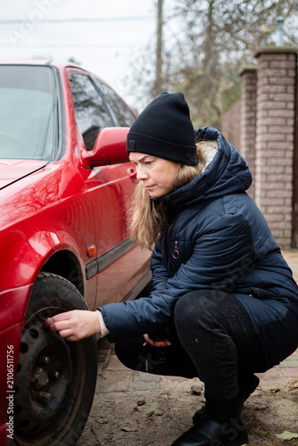 Young Woman in Winter Jacket Inspecting or Changing a Flat Tire on a Red Car Outdoors