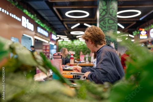 Young person writing notes at wooden cafeteria table beside meal tray and reusable bottle in casual indoor dining space with relaxed focused posture and everyday student atmosphere