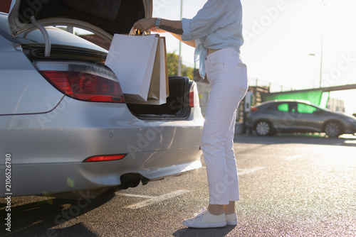 Woman placing white and brown shopping bags into car trunk after successful retail therapy, enjoying black friday discounts and consumerism in a parking lot