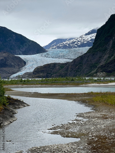 Foto Mendenhall Glacier flowing between rugged mountains with braided glacial streams in the foreground under an overcast sky, Juneau, Alaska, USA