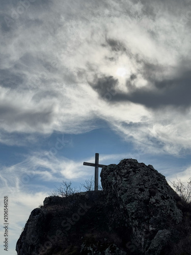 Wooden cross on rocky hilltop under cloudy sky with sunlight breaking through.