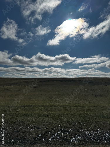 Sunny sky with scattered clouds over open green field and patches of snow.
