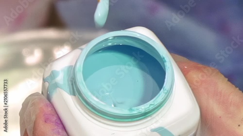 A gloved hand gently dips a golden-bristled brush into a pot of vivid aqua paint. The smooth, liquid color swirls as the brush is immersed, ready for artistic creation. A perfect moment.