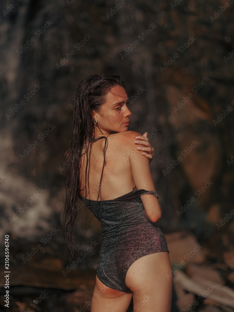 Fototapeta premium Beautiful woman in glitter swimsuit standing back to camera looking shoulder near waterfall