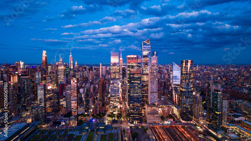 Hudson Yards skyline under layered clouds at blue hour. Aerial view shows 30 Hudson Yards, the Vessel, rail yards and illuminated glass towers beneath textured clouds.