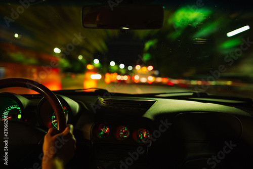 Interior view from behind the steering wheel of a car driving through a dark city street at night with blurry lights.