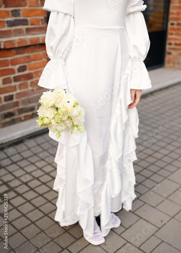 Obraz na plátně Bride wearing a chic long-sleeved white ruffled wedding gown holding a delicate bouquet of white flowers while standing outside near a brick wall