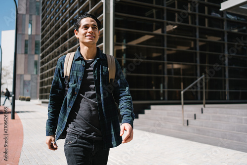 Young hispanic student walking outdoors near modern office university building in urban city carrying backpack
