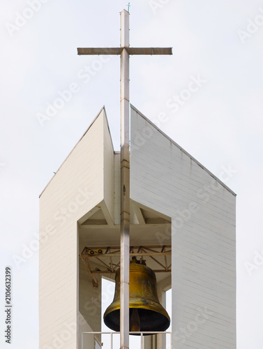 Canvas Print A low angle shot of a modern architectural church bell tower featuring a large brass bell and a Christian cross on top against a bright sky background, religion and architecture concept
