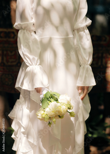 Quadro su tela Bride holding a fresh white flower bouquet while standing outdoors with soft dappled sunlight and leaf shadows falling on her white ruffled dress