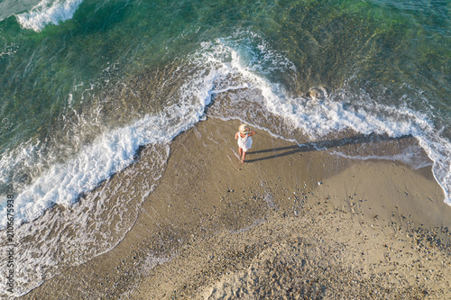 A woman in a white dress and straw hat stands on the shore of a sandy beach. The ocean's surface washes over her feet. View from above, from a drone.