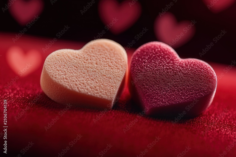 Obraz premium Two porous heart-shaped foam pieces resting on red velvet with soft heart bokeh and warm romantic mood