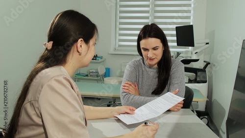 Female doctor asks patient about illness symptoms to fill form in clinic office. Physician listens to woman and holds papers in hospital