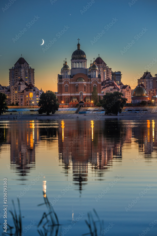 Fototapeta premium Young crescent moon setting after sunset above the Cathedral of the Nativity of Christ on Obolon Embankment in reflected in the calm waters of the Dnipro River