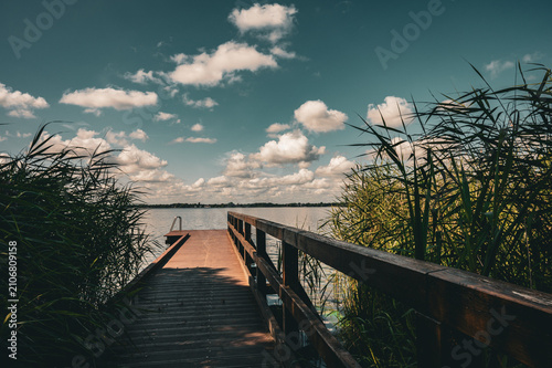 A dark wooden jetty leads into a calm lake surrounded by dense vegetation under a cloudy sky; swimming jetty near Dreibergen on the Bad Zwischenahner Meer, 26160 Bad Zwischenahn, Germany.
