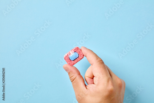 Hand holding single dishwasher detergent tablet on blue background