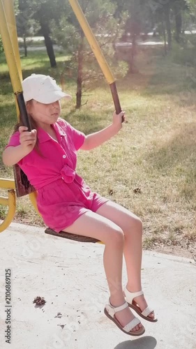 Cute little girl in a white cap and pink clothes enjoys exercise machine on a sunny summer day