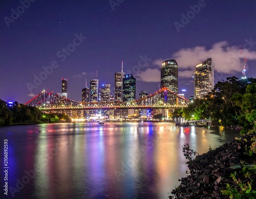 A nighttime cityscape with a bridge over a calm body of water, reflecting vibrant colors, set against a dark sky with a few clouds