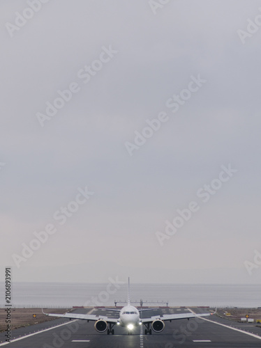 Airplane on runway with lights on, positioned for takeoff, partially cloudy sky visible, near the coast