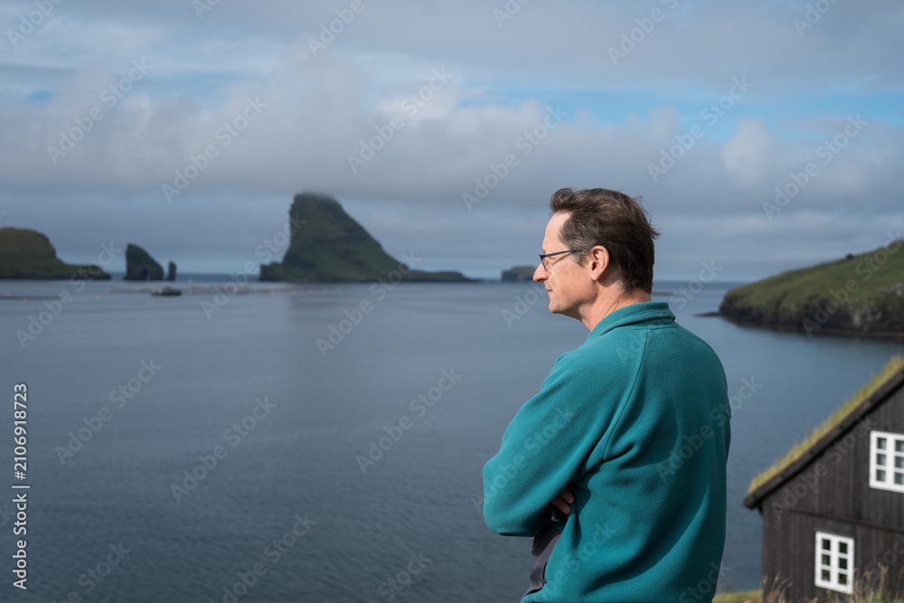 Fototapeta premium Mature Man Over 50 Looking Out at Tindholmur Islet from Bøur Village in Faroe Islands