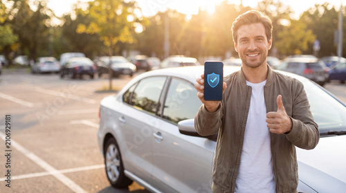 Smiling man holding smartphone with security icon beside sedan in parking lot at sunset