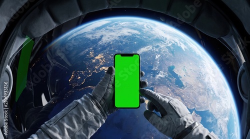 Astronaut man in space suit holding smartphone with green screen. Earth view from international space station. Concept of futuristic mobile communication and technology in outer space exploration.